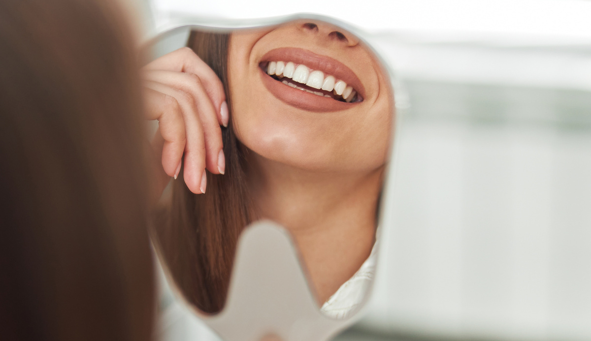Happy young woman smiling checking out her perfect healthy teeth in the mirror close up, at the dentist office